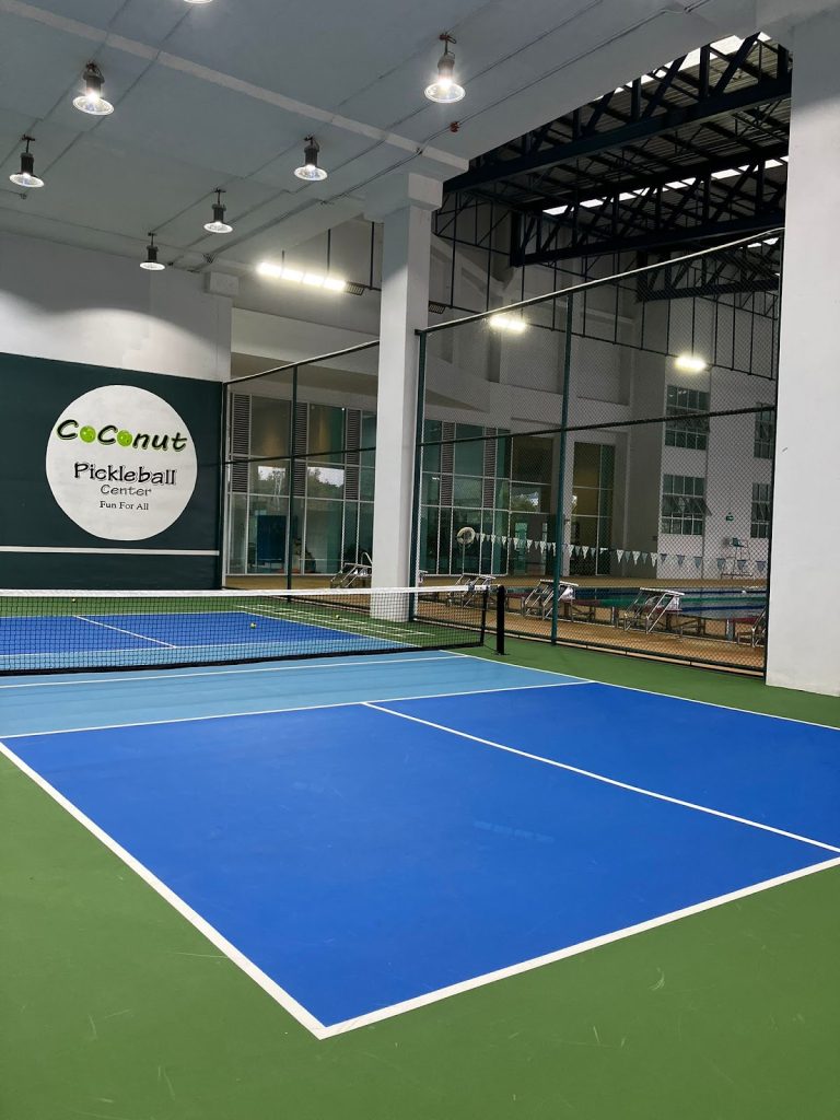 Indoor pickleball court with a blue playing area, green borders, and a net. Large logo on the wall reads "Coconut Pickleball Center." Ceiling lights illuminate the space.