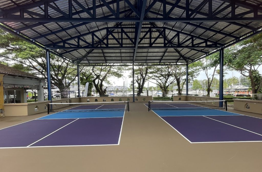 Indoor sports facility with two empty pickleball courts, sheltered by a metal roof. Surrounding area features trees and a marina in the background.