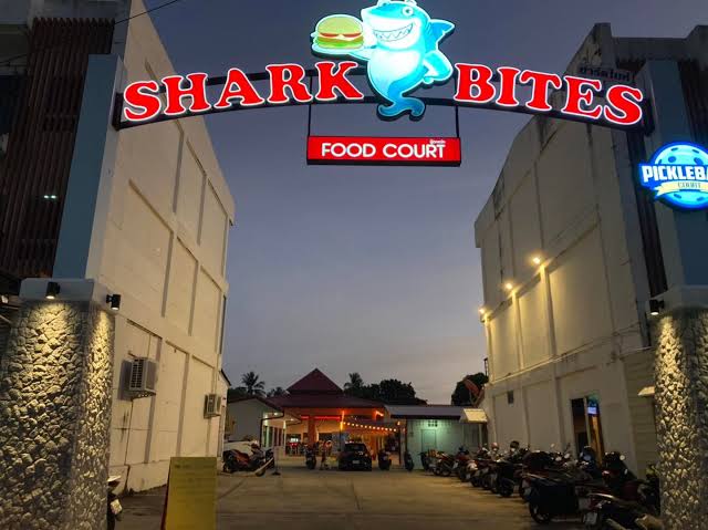 Entrance to Shark Bites Food Court with a smiling shark logo, illuminated sign, and motorcycles parked along the sides.