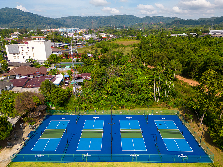 Aerial view of four blue tennis courts surrounded by greenery and buildings, with hills in the background.
